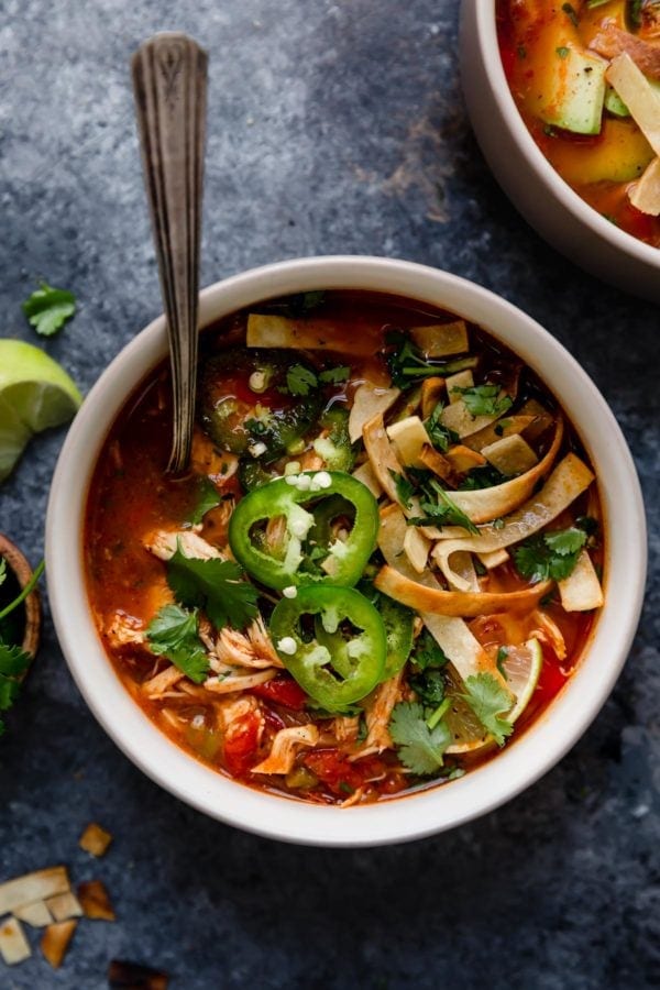 A white bowl on a grey surface filled with Instant Pot Chicken Tortilla-Less Soup that's topped with slices of fresh jalapeños, baked tortilla strips, and a lime wedge. 