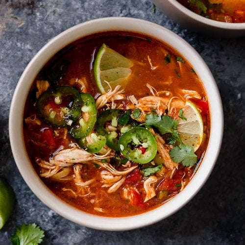 A white bowl on a grey surface filled with Instant Pot Chicken Tortilla-Less Soup that's topped with slices of fresh jalapeños and a lime wedge.