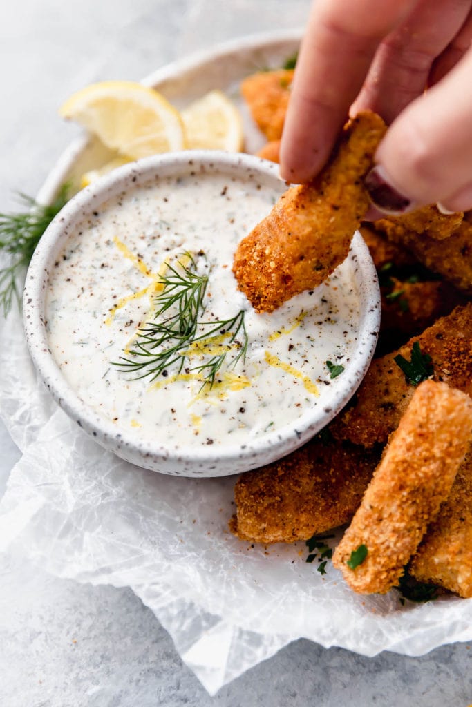 Baked Fish sticks on a serving platter lined with parchment including a bowl of homemade tartar sauce. One of the fish sticks dipped into the tartar. 