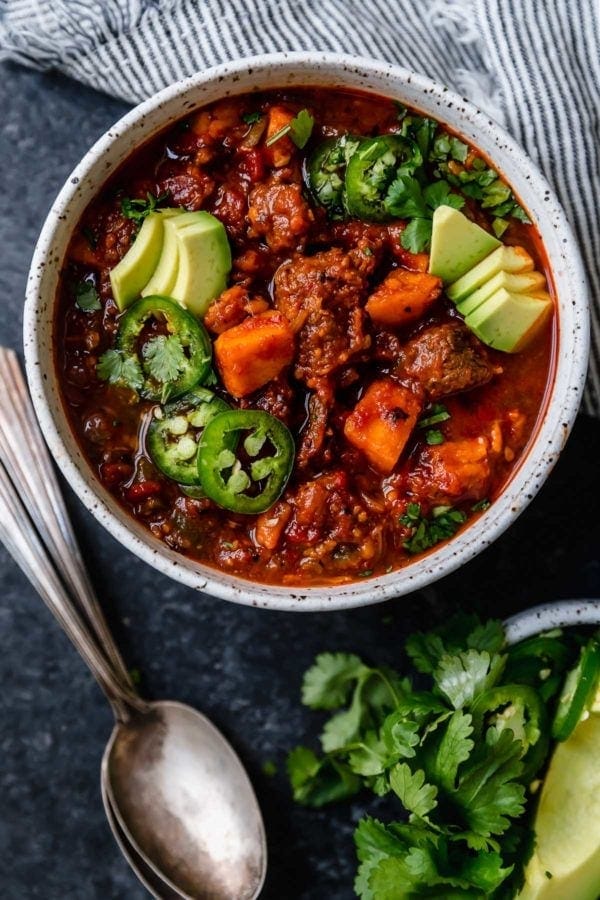 An overhead shot of a white bowl and two silver spoons on a dark surface that's filled with Slow Cooker Chunky Beef Chili with Sweet Potatoes and topped with sliced jalapeños, avocado, and fresh cilantro. 