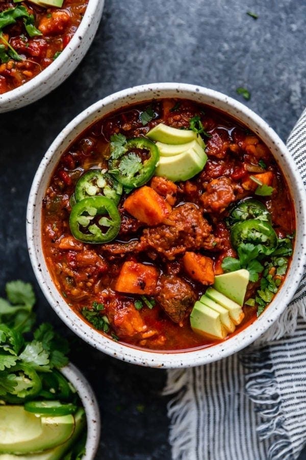 A white bowl on a dark surface that's filled with Slow Cooker Chunky Beef Chili with Sweet Potatoes and topped with sliced jalapeños, avocado, and fresh cilantro. 