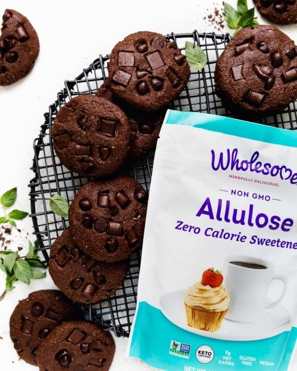 A pile of Low Sugar Double Chocolate Mint Cookies on a round, black cooling rack next to a bag of Wholesome Allulose Zero-Calorie Granulated Sweetener. 