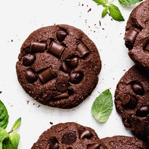Low Sugar Double Chocolate Mint Cookies studded with chocolate chips and chunks on a white surface surrounded by fresh mint leaves.