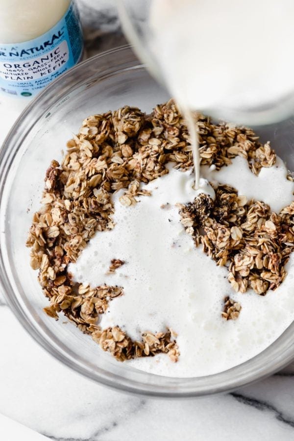 Kalona Supernatural Organic Plain Kefir is being poured from a glass measuring cup into a bowl filled with rolled oats to make Mixed Berry Baked Oatmeal.