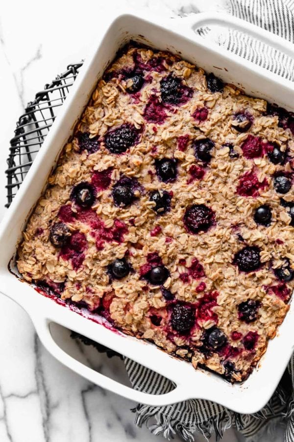 A square white dish filled with Mixed Berry Baked Oatmeal sits atop a wire cooling rack on a marble surface. 