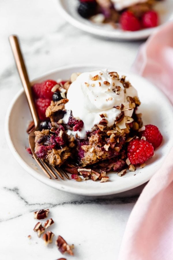 A small white plate holds a serving of Mixed Berry Baked Oatmeal topped with whipped cream and sprinkled with nuts. 