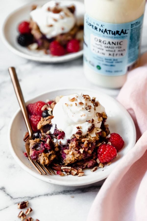 A small white dish holds a serving of Mixed Berry Baked Oatmeal that's topped with whipped cream and sprinkled with toasted nuts ready to be served with a gold fork. In the background, a bottle of Kalona Supernatural Organic Plain Kefir is positioned next to another serving of Mixed Berry Baked Oatmeal. 