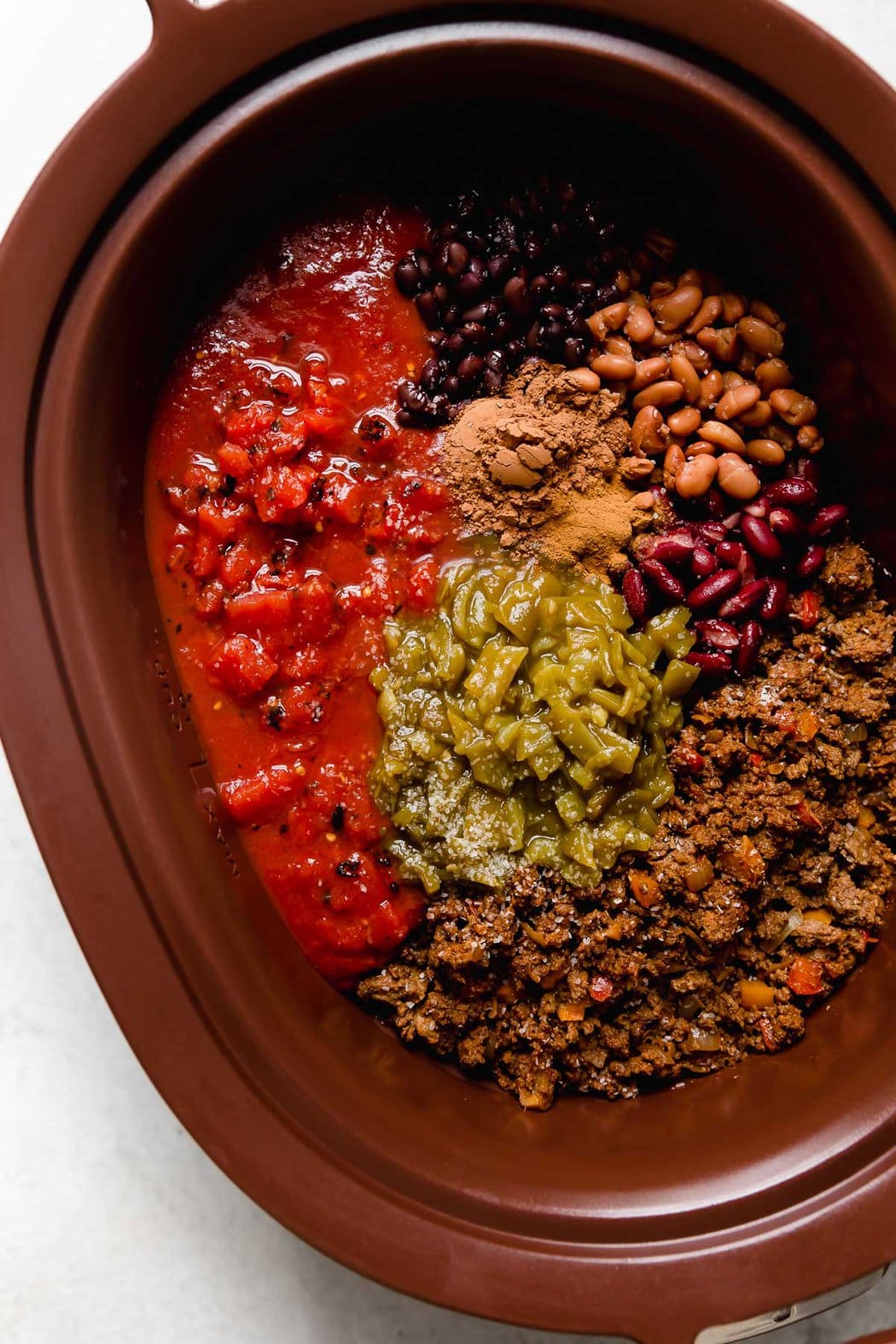 Overhead view dark brown slow cooker filled with all ingredients for beef and bean chili ready to be stirred together