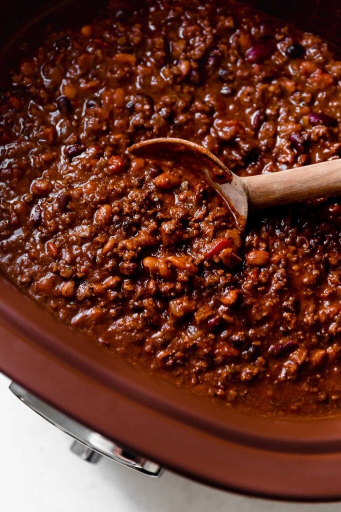 Wooden spoon scooping up spoonful of slow cooker beef and bean chili from crock pot