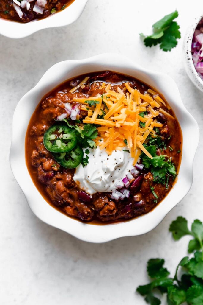 Overhead view serving of slow cooker beef and bean chili in white bowl, topped with cheddar shredded cheese, sour cream and jalapeno slices.