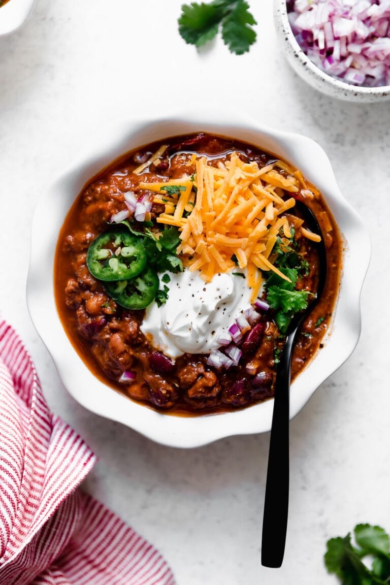 Overhead view of slow cooker beef and bean chili in white bowl topped with cheese, sour cream, jalapenos and chopped red onions. 