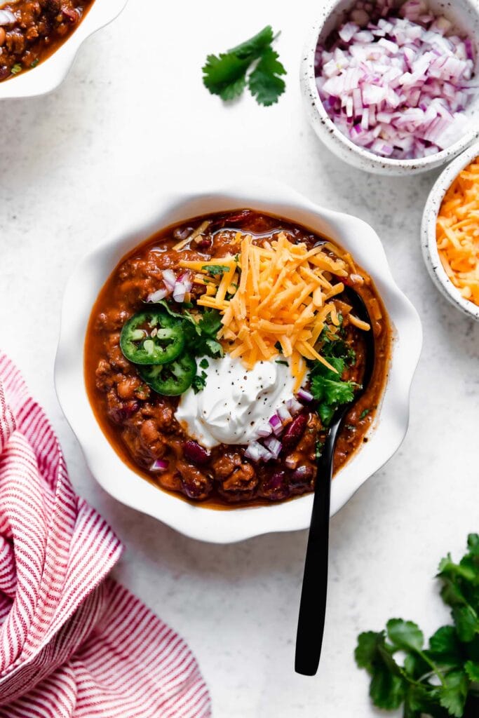 Overhead view white bowl filled with slow cooker beef and bean chili topped with shredded cheddar cheese, sour cream and jalapeno slices.