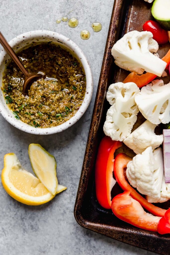 Overhead view sheet pan with raw vegetables, small bowl with chicken marinade for Mediterranean chicken to the side of sheet pan