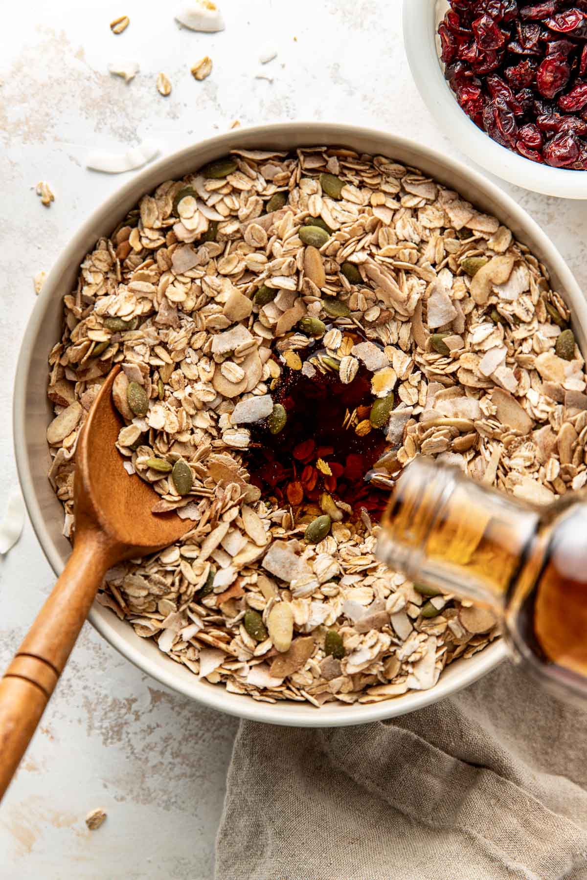 Overhead view of a bowl of dry ingredients for Homemade Granola with maple syrup being poured into the mixture. 