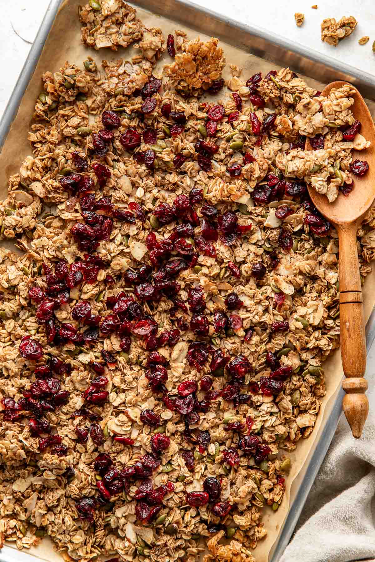 Overhead view of a freshly baked batch of granola on a sheet pan topped with dried cherries and a wooden spoon on the side. 