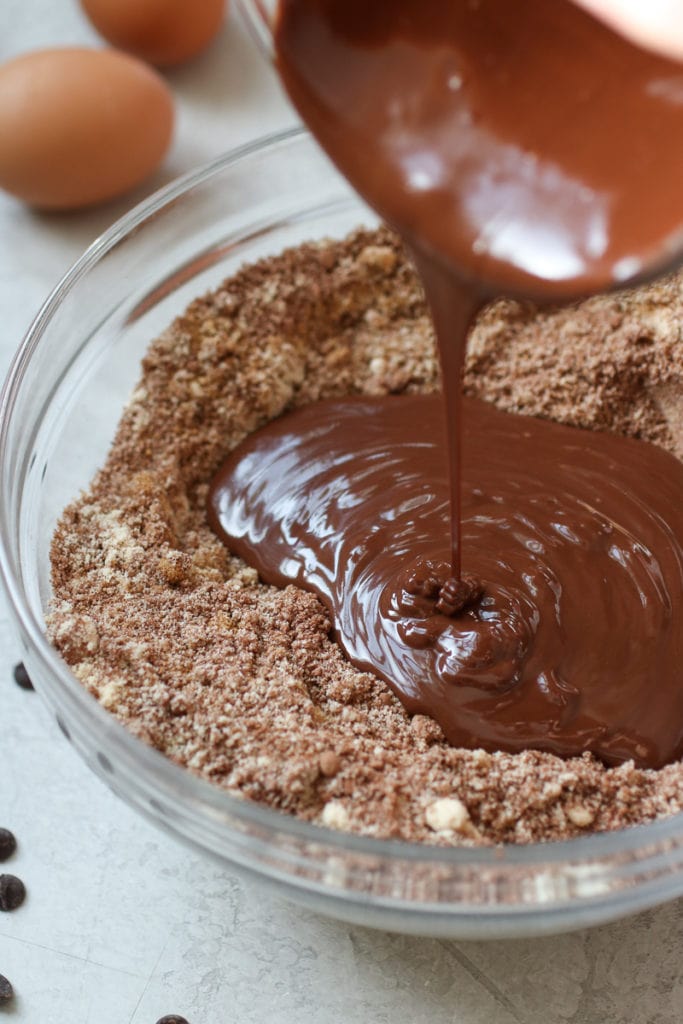 Photo of melted chocolate being poured into a clear bowl with dry ingredients for Chocolate Peanut Butter Protein Cookies. 