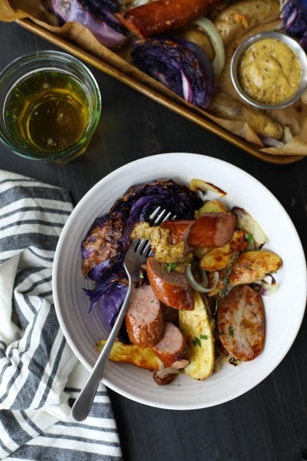 Overhead view of a portion of Sheet Pan Kielbasa, Cabbage and Potatoes served in a white bowl with a glass of lager. 