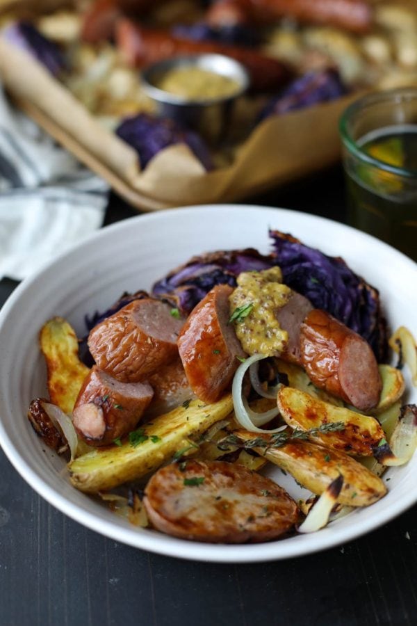 A white bowl piled with roasted potatoes, cabbage, onions, and kielbasa with a generous dollop of grainy mustard.