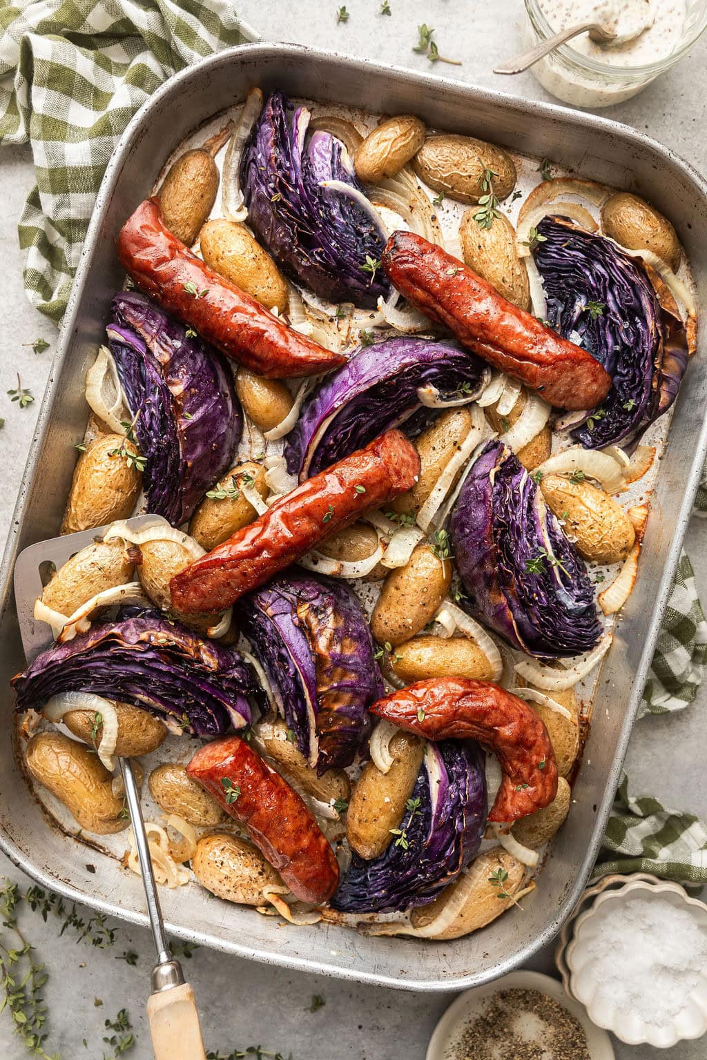 Overhead view of a sheet pan of kielbasa and potatoes with wedges of purple cabbage topped with fresh herbs and black pepper.