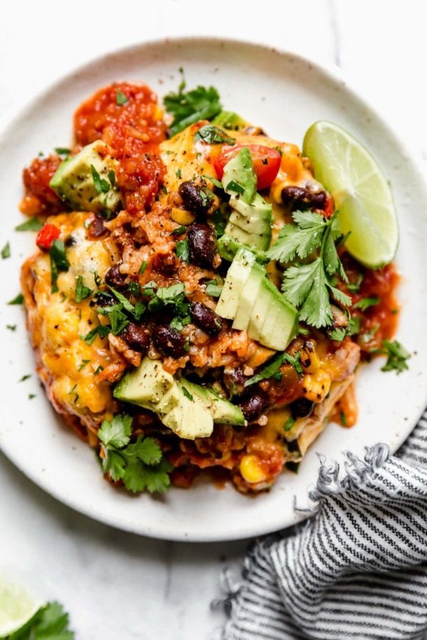 White plate with serving of taco casserole topped with avocado and fresh lime slices.