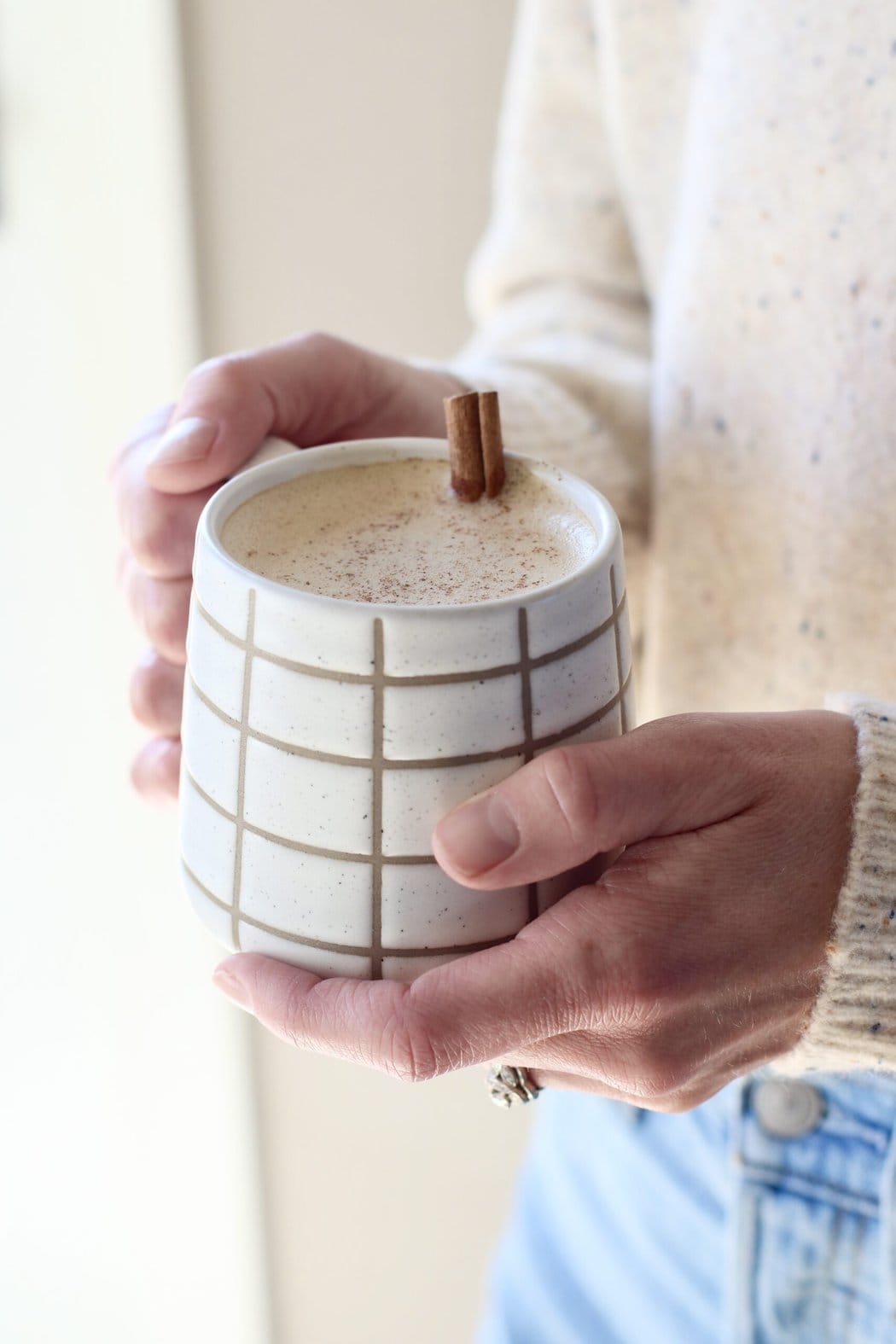 Close up view of hands holding a white coffee mug of Cinnamon Coconut Latte topped with cinnamon sprinkles on top. 