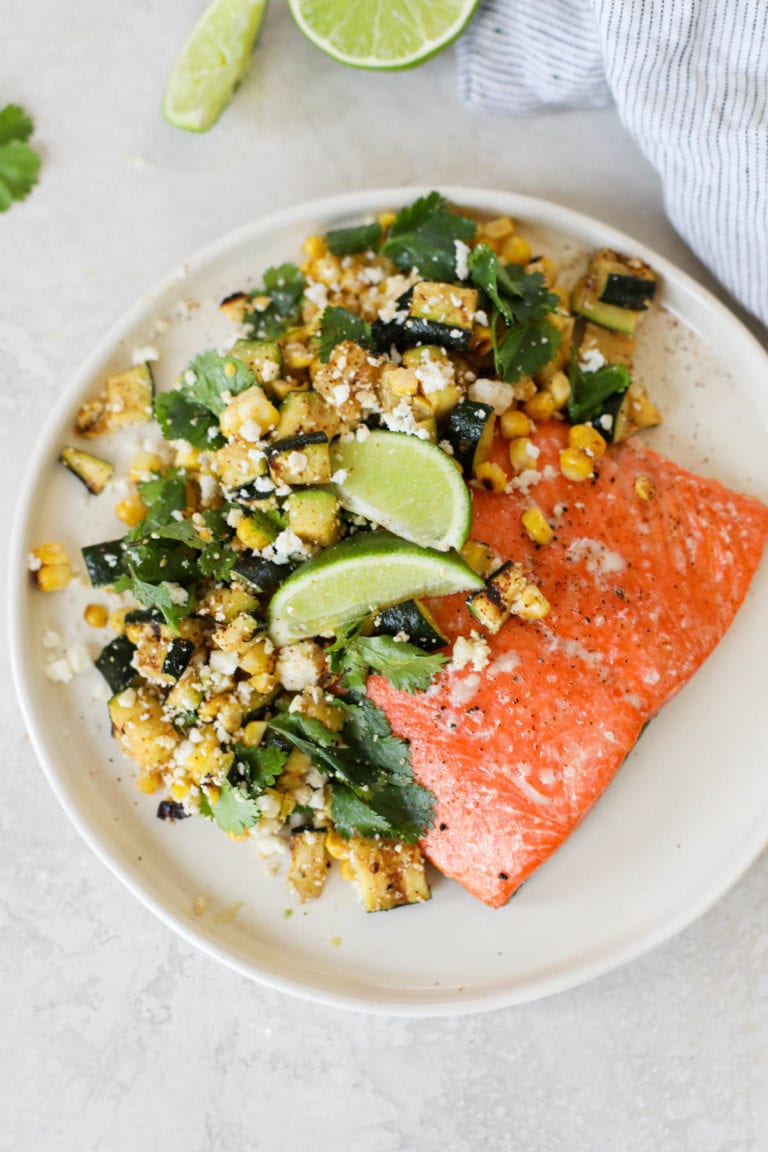 Overhead view of a plate of grilled salmon with elote-style vegetables garnished with lime wedges. 