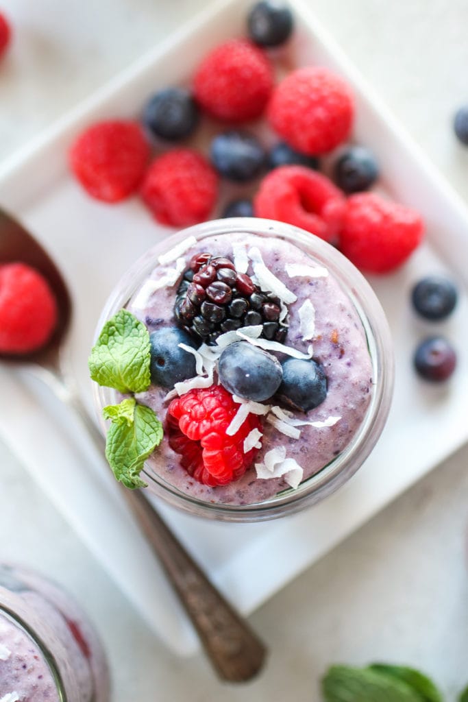 Overhead, close-up photo of Mixed Berry Protein Chia Pudding on a white square plate. Berries onto to the pudding with coconut flakes and mint sprig. Berries and a spoon also on the plate.