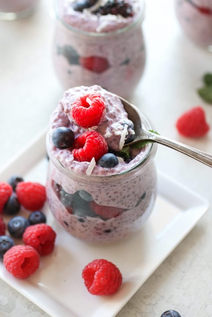 Photo of Mixed Berry Protein Chia Pudding in a glass jar layered with berries and garnished with a sprig of mint. Spoon in the jar holding a bite-size amount of the pudding.