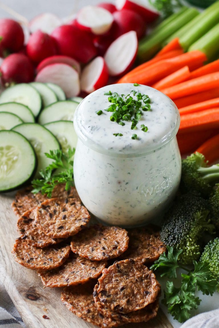 Creamy homemade ranch dressing in small glass jar on tray surrounded by fresh cut vegetables and crackers