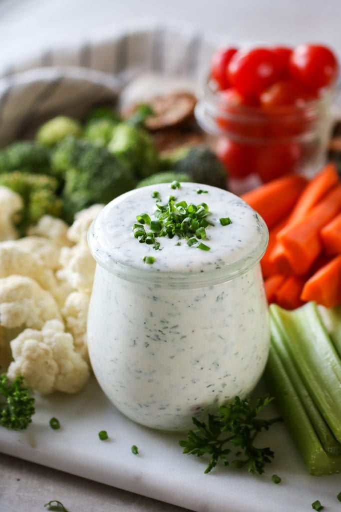 Photo of Healthy Homemade Ranch Dressing in a mason jar topped with fresh chives and surrounded by fresh cut vegetables.