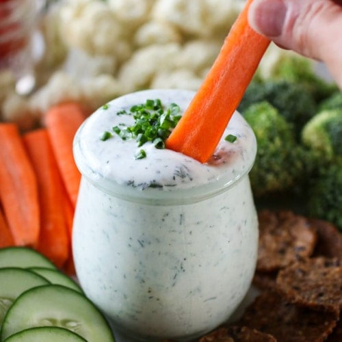 Small jar of homemade ranch dressing on cutting board surrounded by fresh cut vegetables