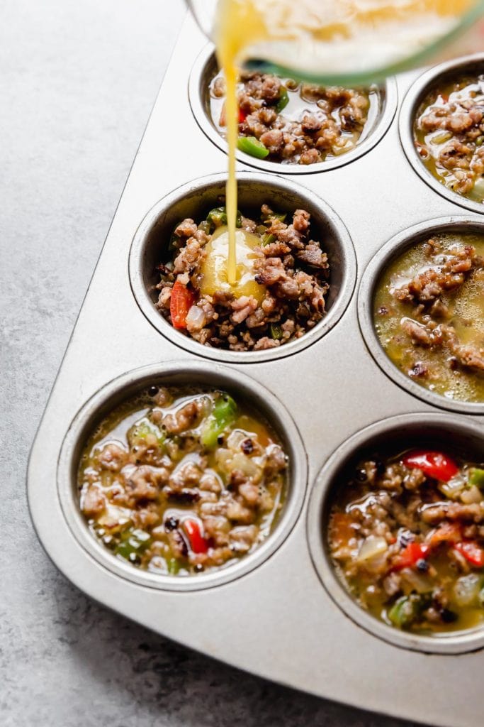 Prep photo of Sausage Hash Brown Egg Muffins being made. Whisk eggs being poured in a muffin well that's filled with sausage, hash browns and green and red peppers.