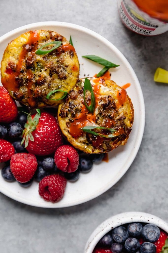 Photo of two Sausage Hash Brown Egg Muffins on a white with berries. Garnished with green onion and drizzled with hot sauce.