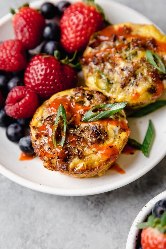 Photo of two Sausage Hash Brown Egg Muffins on a white with berries. Garnished with green onion and drizzled with hot sauce.