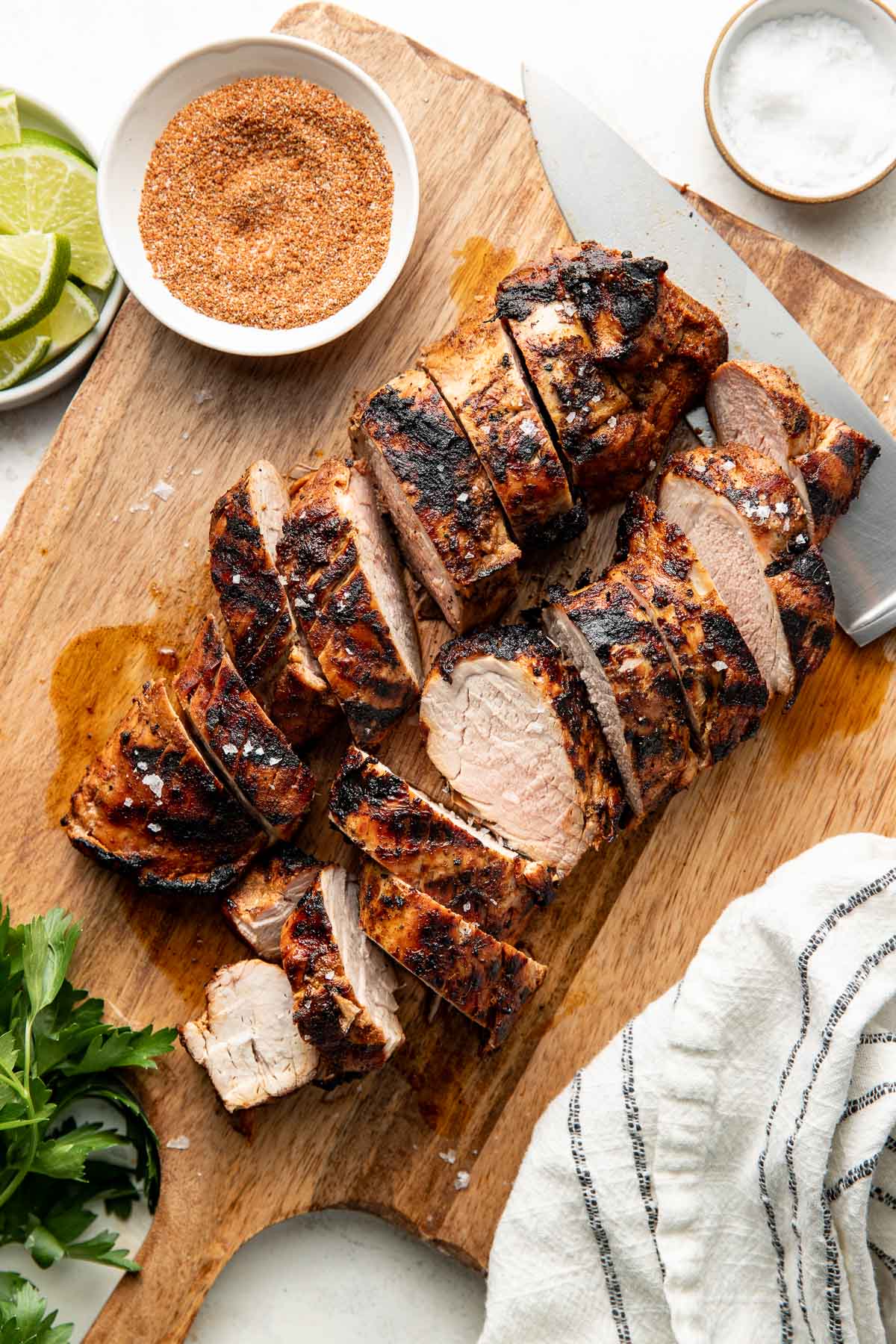 Overhead view of sliced grilled pork tenderloin sprinkled with flaky sea salt on a wooden cutting board with a knife and striped towel.