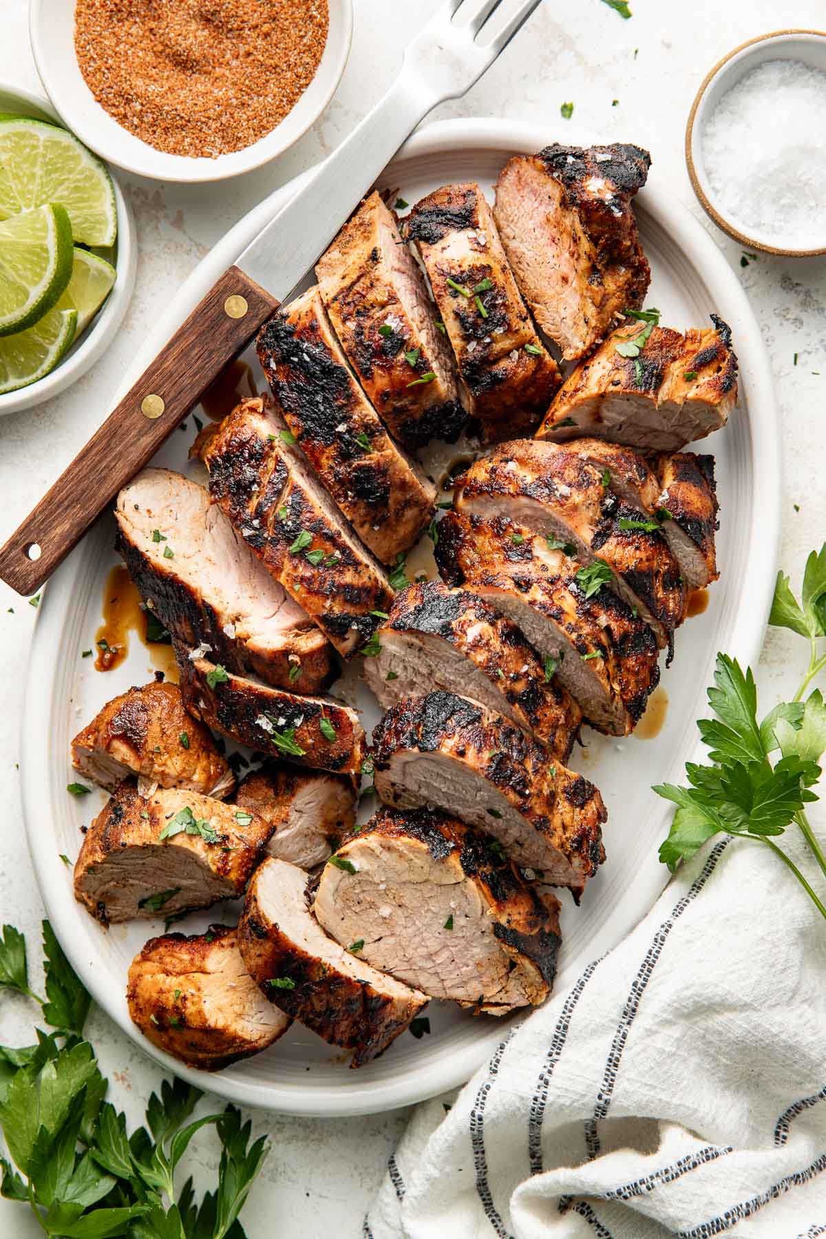 Perfectly grilled and sliced pork tenderloins arranged on a white oval platter surrounded by fresh parsley, limes and red spice mixture in a white bowl.
