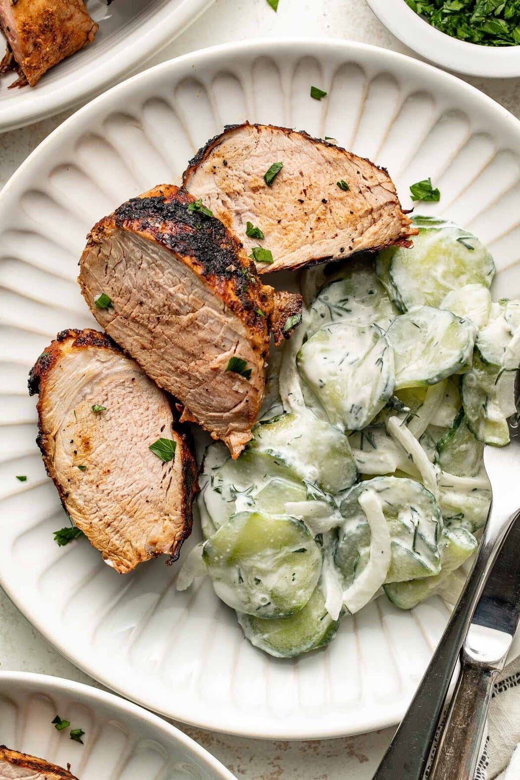 Closeup overhead of a white plate with silver utensils and sliced pork with a creamy cucumber salad.