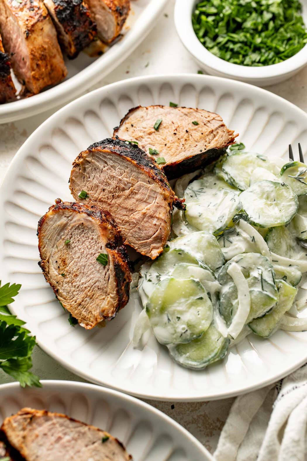 Three slices of tender grilled pork loin on a white plate with creamy cucumber salad.