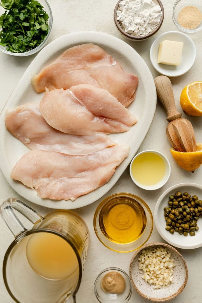 An overhead view of ingredients for making lemon chicken piccata including chicken, flour, butter, lemons, capers, olive oil, chicken broth, garlic and dijon mustard. 
