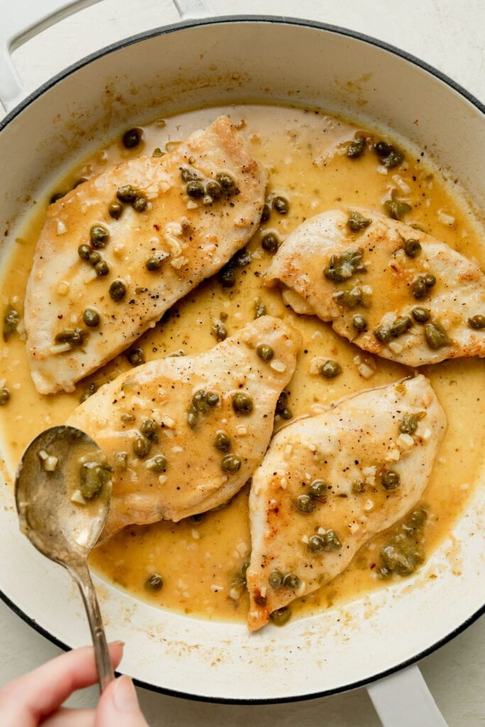 A hand spooning a lightly thickened lemony sauce with garlic and capers over chicken breasts in a white skillet. 