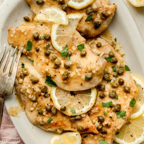 A large platter of chicken piccata cutlets garnished with capers, parsley and lemons.