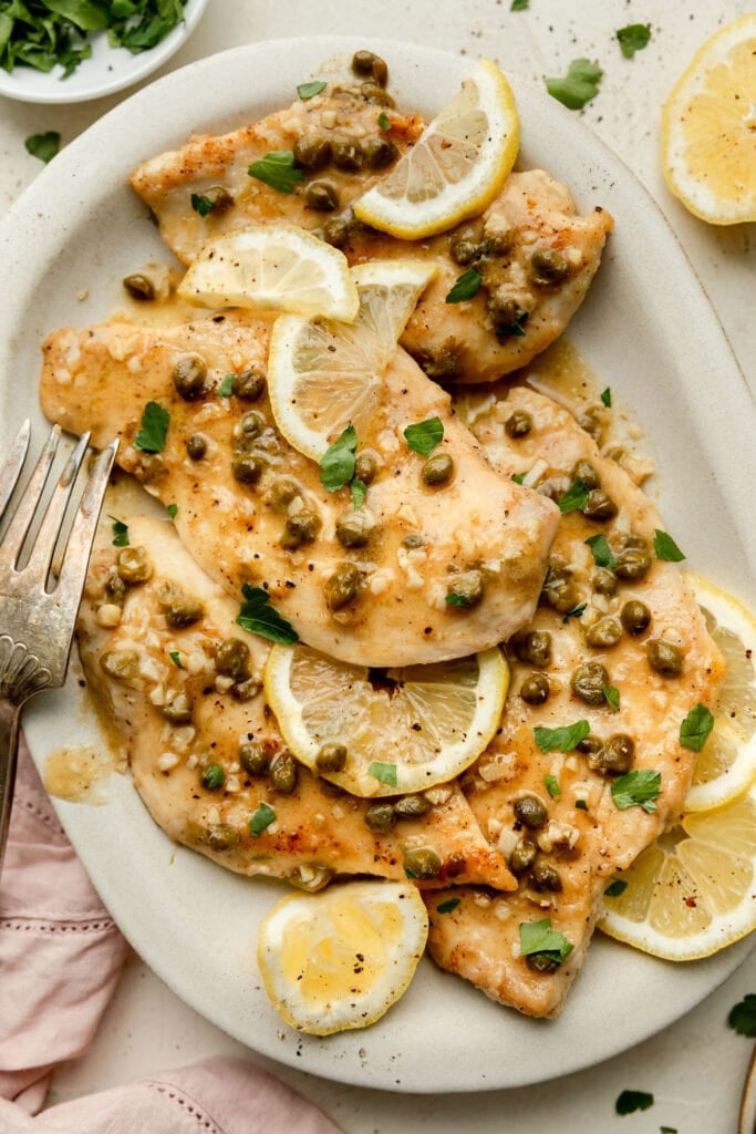 A large platter of chicken piccata cutlets garnished with capers, parsley and lemons.