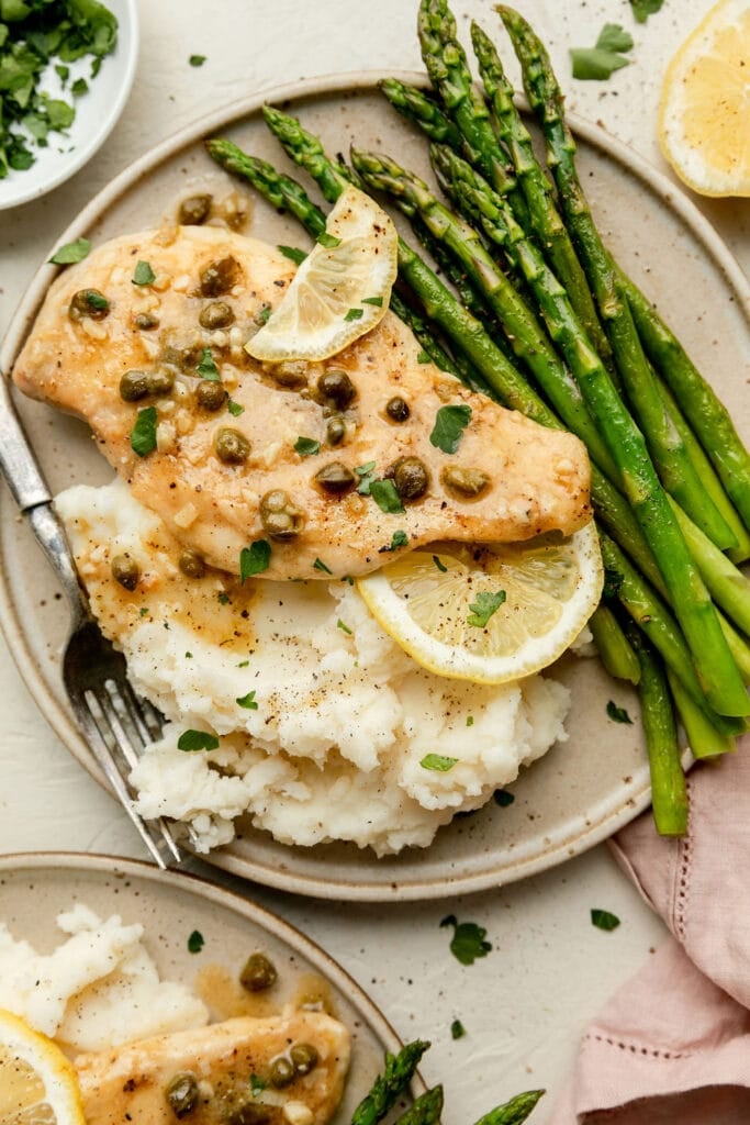 A plate with lemon chicken piccata, mashed potatoes and fresh asparagus garnished with a lemon slice. 