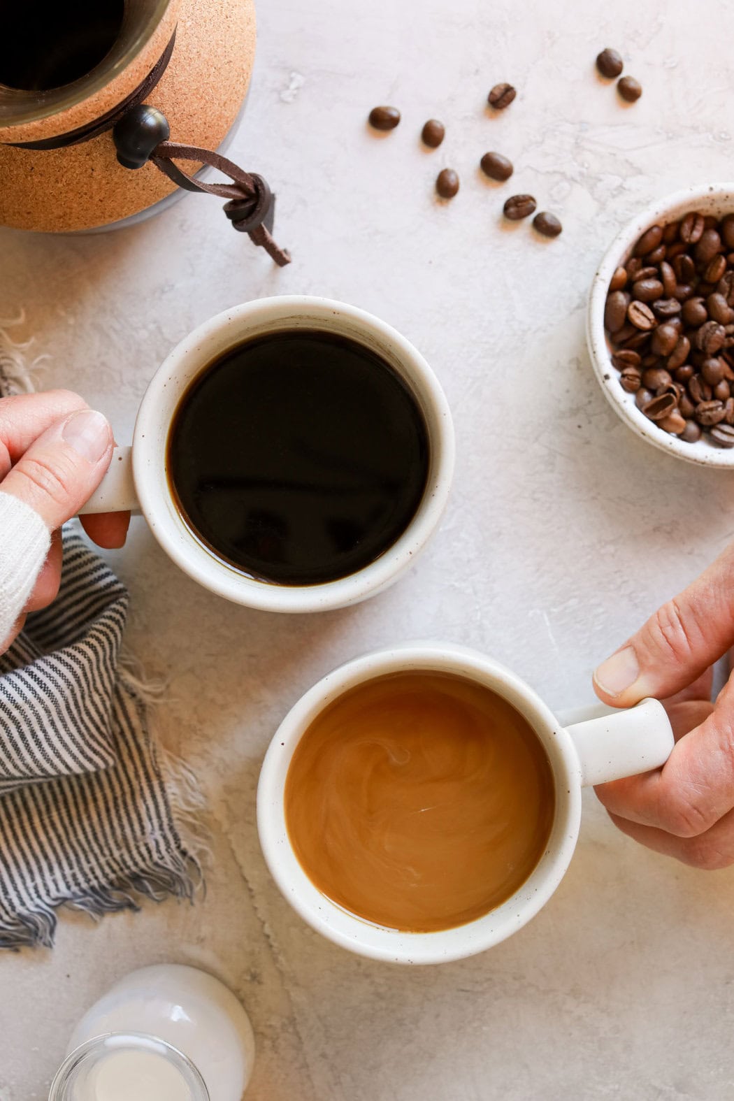 Overhead view of hands holding coffee mug handles, one filled with black coffee and the other filled with coffee and cream. 