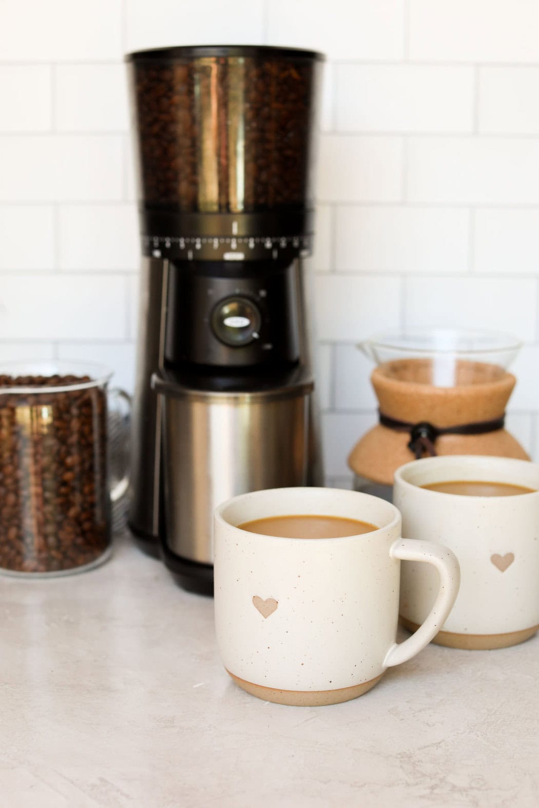 Two white coffee mugs filled with coffee next to a coffee grinder and whole coffee beans. 