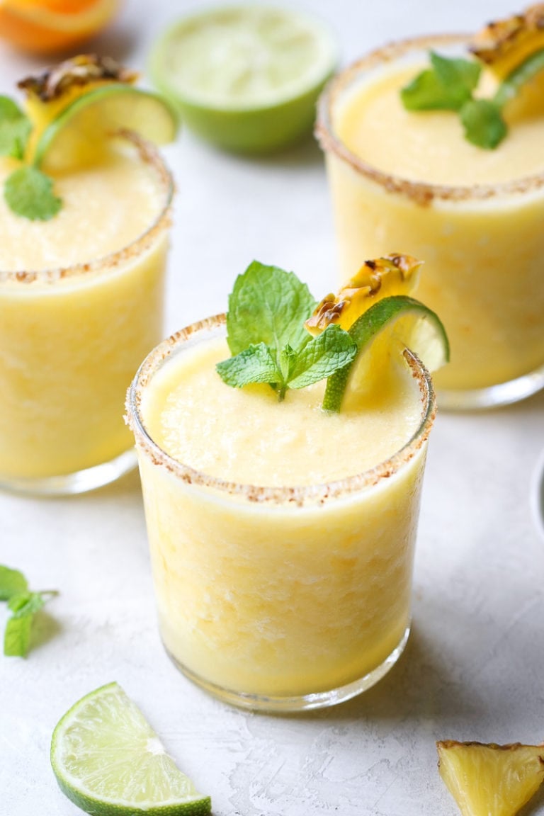 Overhead view of several frozen pineapple margaritas topped with fresh mint leaves and lime wedges. 