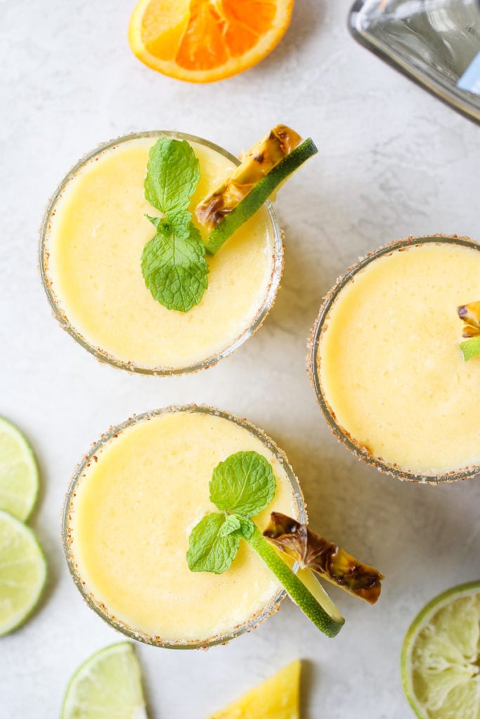 Overhead photo of Frozen Pineapple Margaritas - three servings garnished with a fresh mint leaf, lime slice and pineapple wedge.