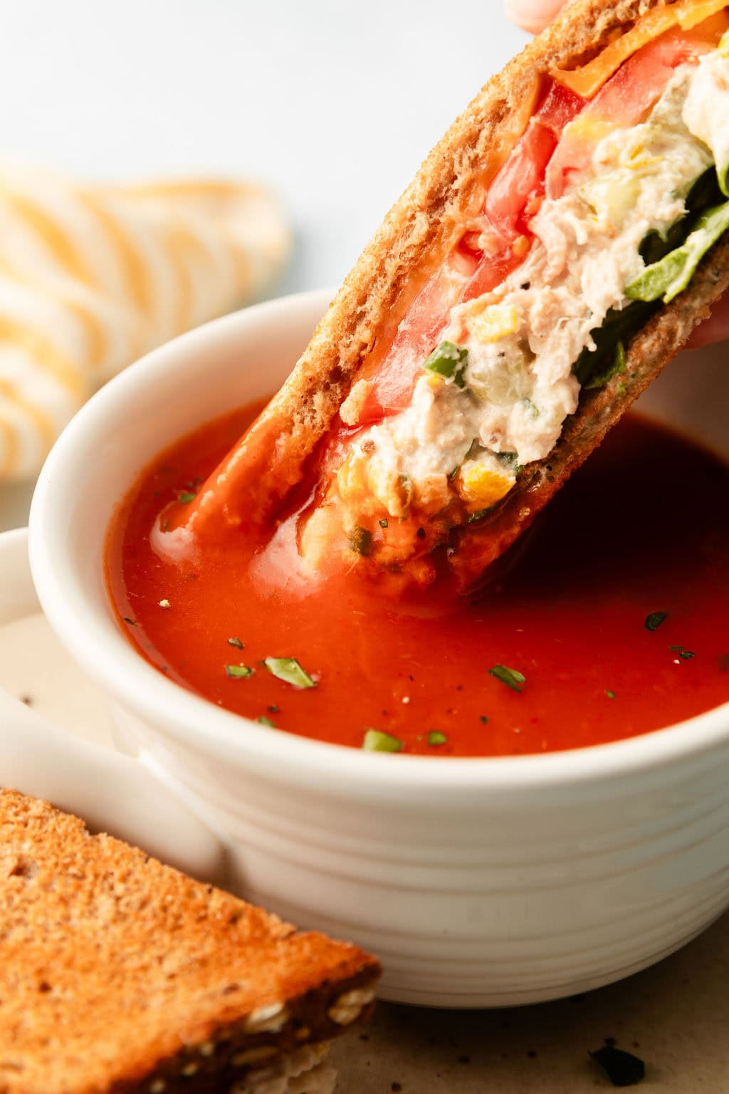 Close up view of a tuna melt being dunked into a bowl of tomato soup.