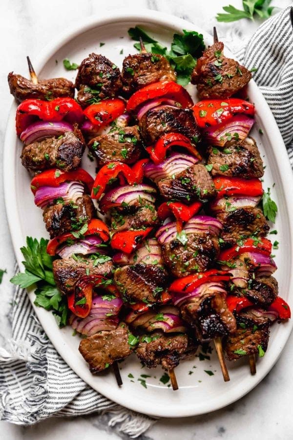 Several grilled steak kebabs on skewers with red peppers and purple onions, on a white platter