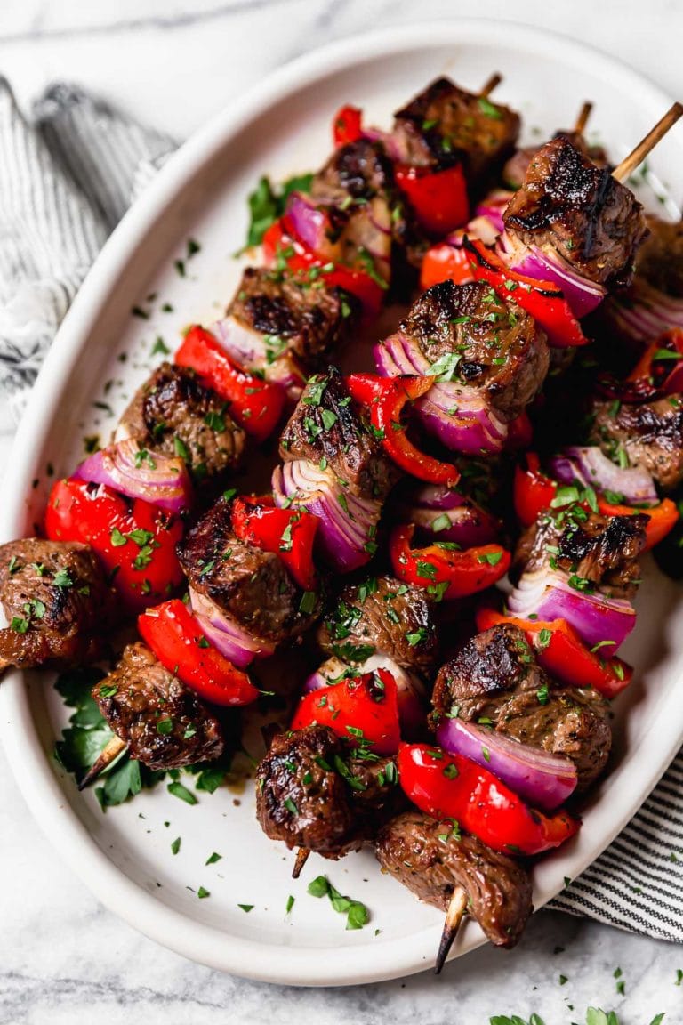 Grilled Steak Kebabs on a white plate topped with fresh herbs and accompanied by red pepper and red onions on the skewers.